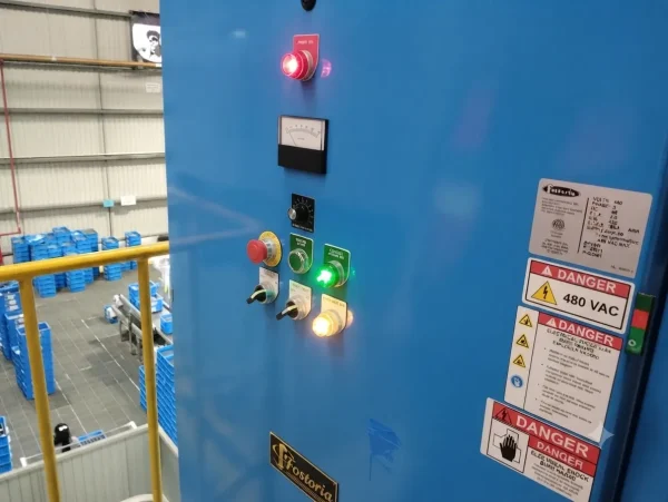 Tablero de control industrial de color azul con interruptores, luces indicadoras, medidores y etiquetas de advertencia de alto voltaje 480 VAC en una planta de manufactura.